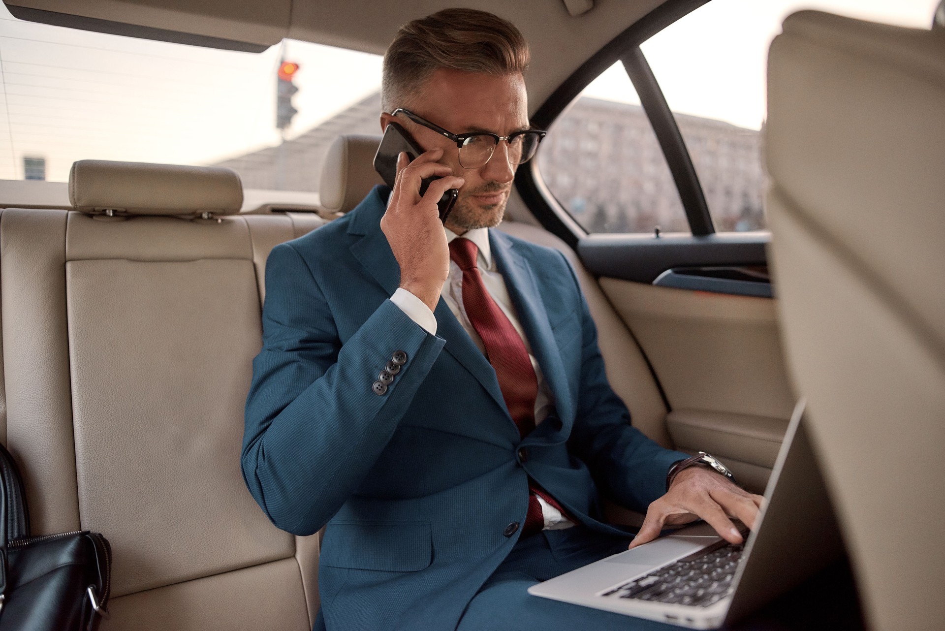 Discutindo detalhes do projeto. Homem de negócios maduro confiável e à moda no terno cheio que trabalha em seu portátil e que fala no telefone com sócio de negócio ao sentar-se no carro
